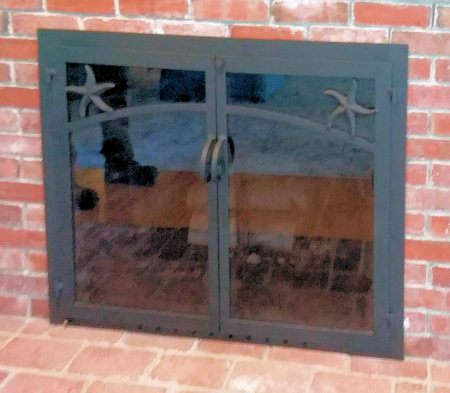 Cotuit fireplace doors all black finish with starfish motif. twin doors standard forged handles and smoked glass. Comes with slide mesh